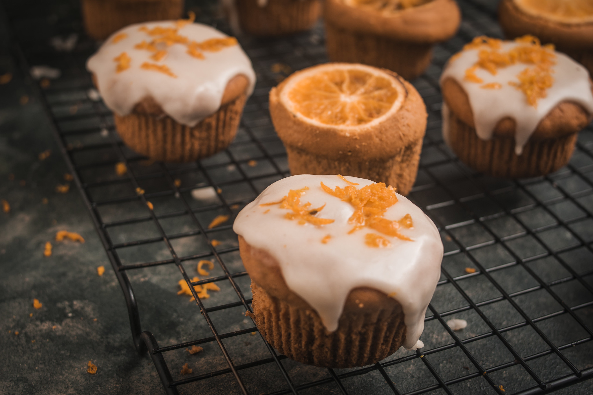 Close-up of iced blood orange cupcakes on a cooling rack, finished with a thin white glaze and scattered fresh orange zest.
